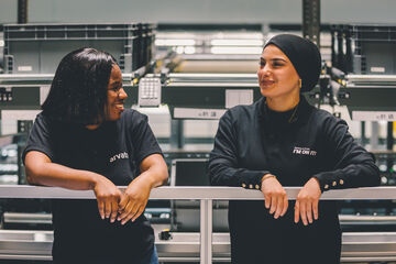 Zwei lächelnde weibliche Angestellte in schwarzen Arvato-T-Shirts in einer Lagerhalle