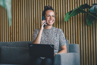 a woman sits on a gray couch with a laptop on her lap and is on the phone