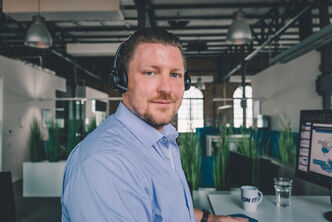 An Arvato customer service agent with headset at a desk, looking into the camera
