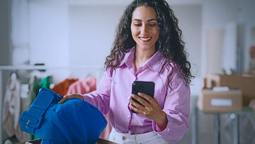 Smiling woman in purple blouse holds blue cloth pants in one hand and looks into smartphone in her other hand