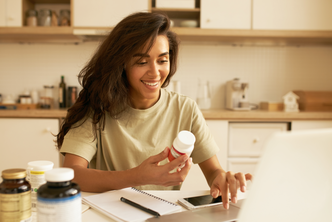 a woman smiles while typing on her laptop and holding a medicine box in her right hand