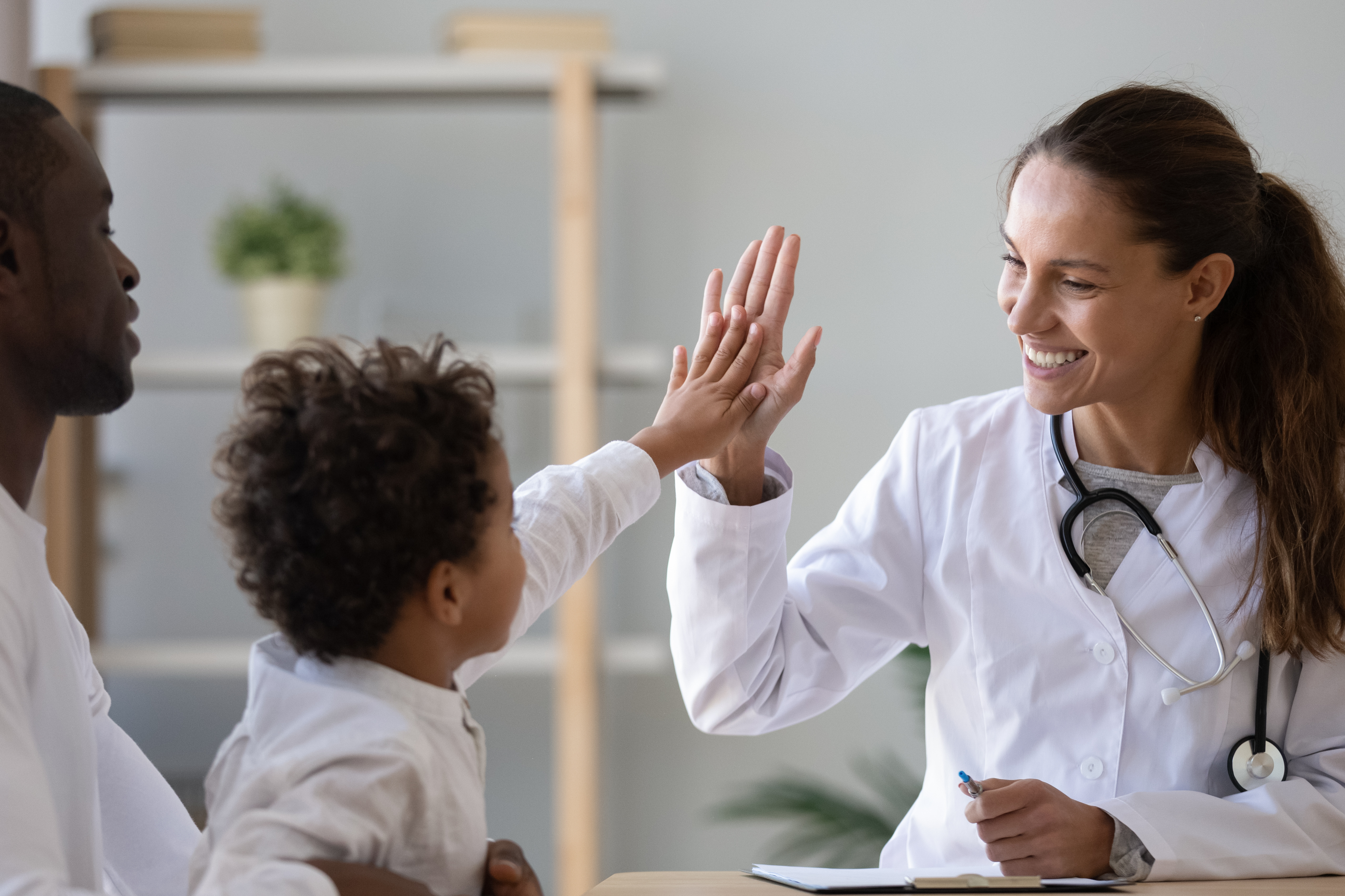 ein kleiner Junge auf dem Arm seines Vaters gibt einer lächelnden Kinderärztin ein High Five